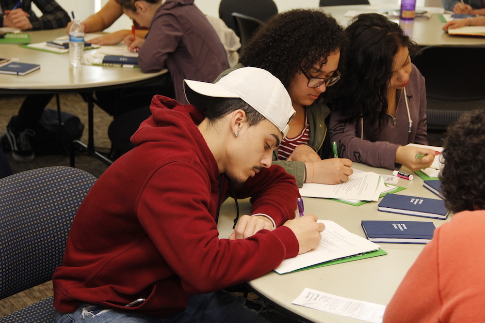 A male student wearing a backwards cap writes in a notebook